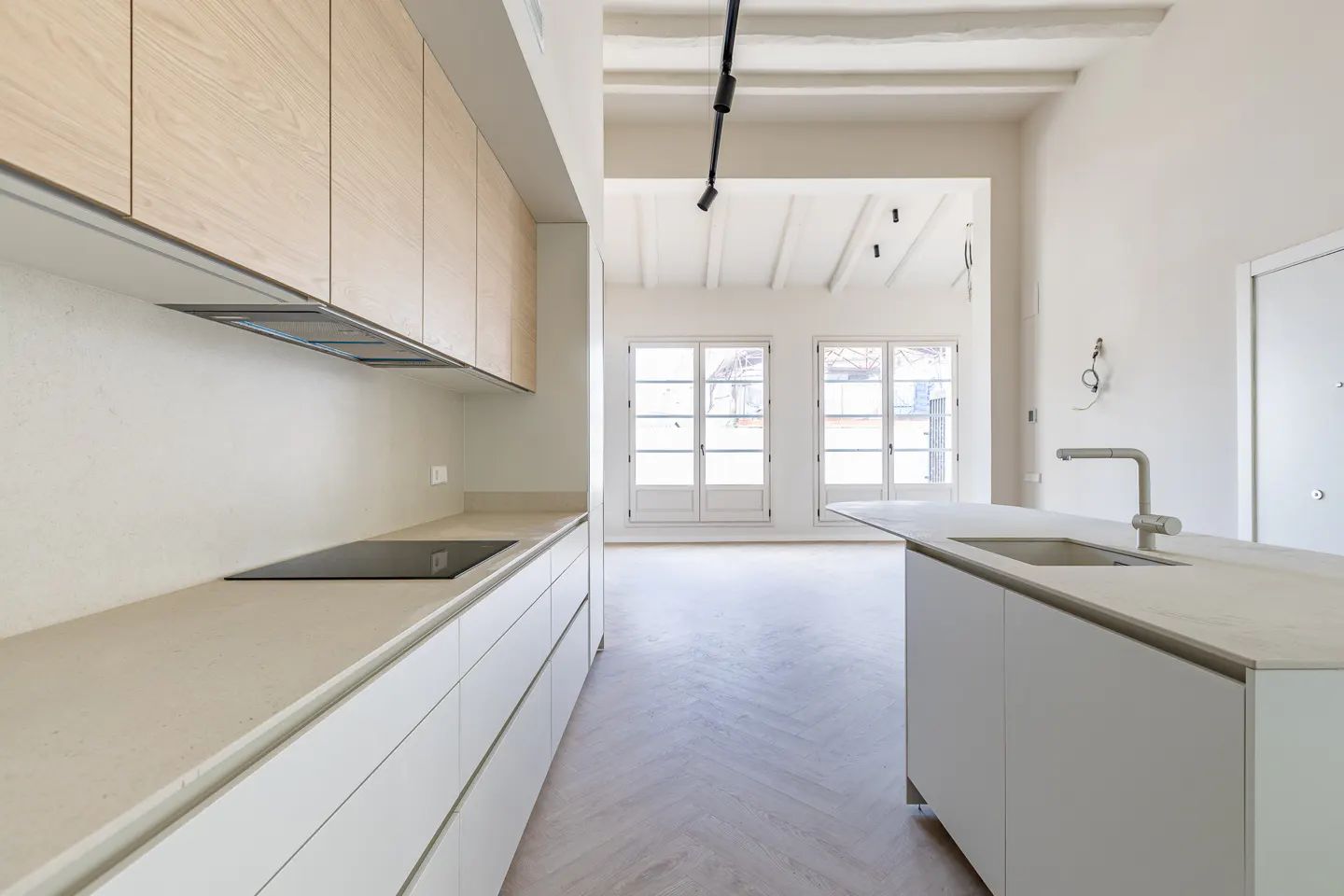 A modern kitchen with light wood cabinets, white countertops, and a central island with a sink. Two windows are in the background.