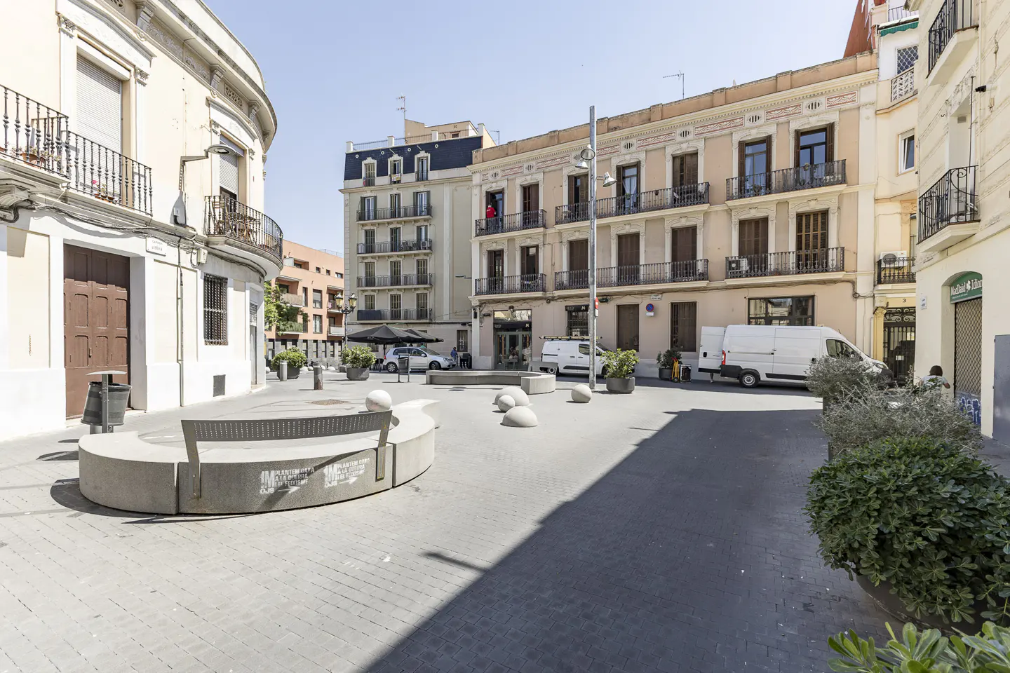 A sunny plaza with stone benches, surrounded by multi-story buildings with balconies and parked white vans.