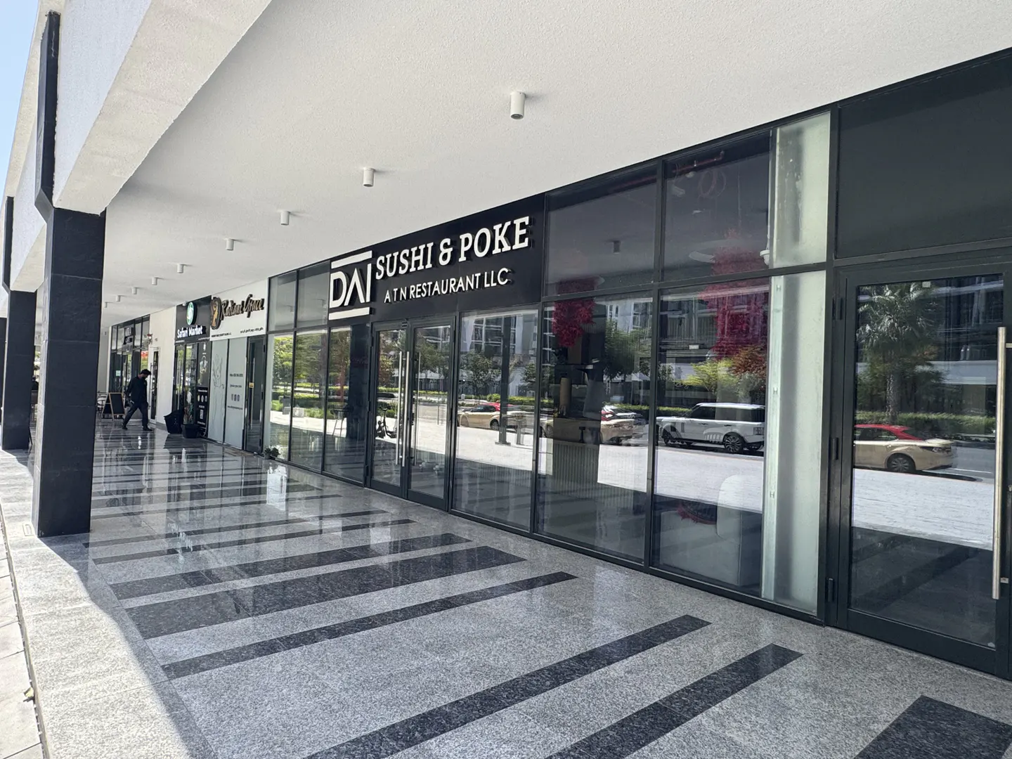 Exterior view of a commercial building with "DAI Sushi & Poke" restaurant. The building has large glass windows and a gray tiled walkway.