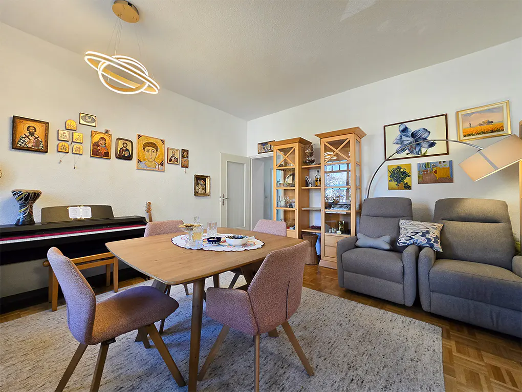 Living room with a piano, dining table with pink chairs, and gray reclining sofas. Paintings and icons decorate the walls.