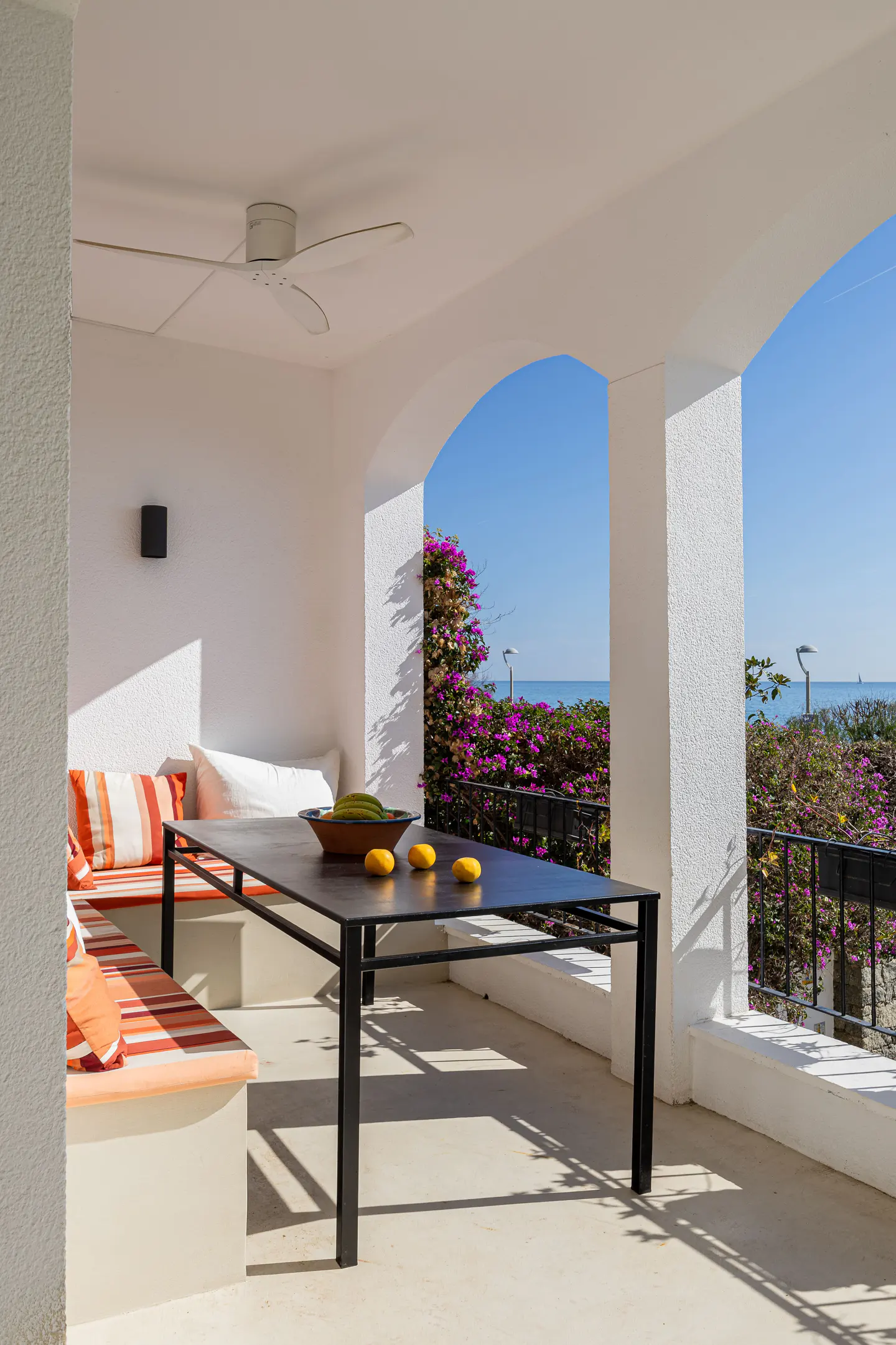 Covered patio with a black dining table, built-in seating with orange striped cushions, and a view of the ocean and flowers.