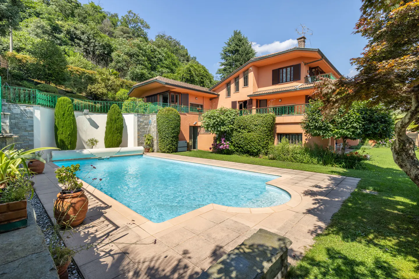 A sunny view of a peach-colored house with a blue pool, green lawn, and trees.