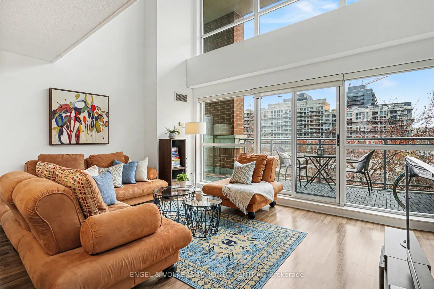 Bright living room with brown sofas, blue rug, and balcony overlooking city buildings. Modern art hangs on the white wall.