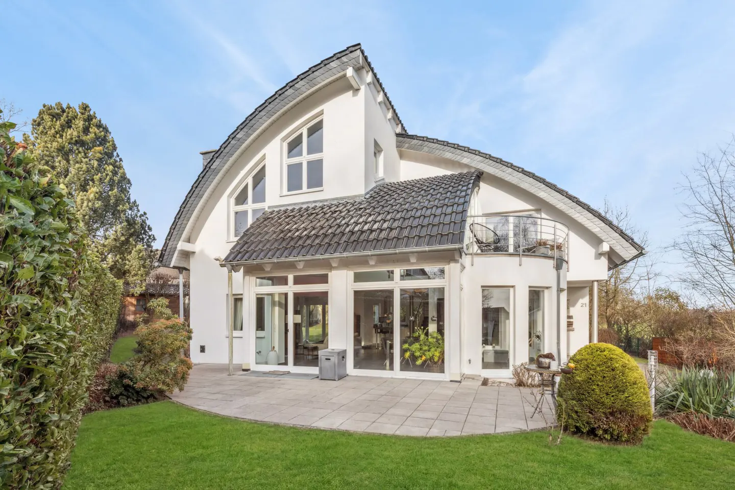 Rear view of a modern white house with a curved roof, large windows, and a stone patio surrounded by green lawn and landscaping.