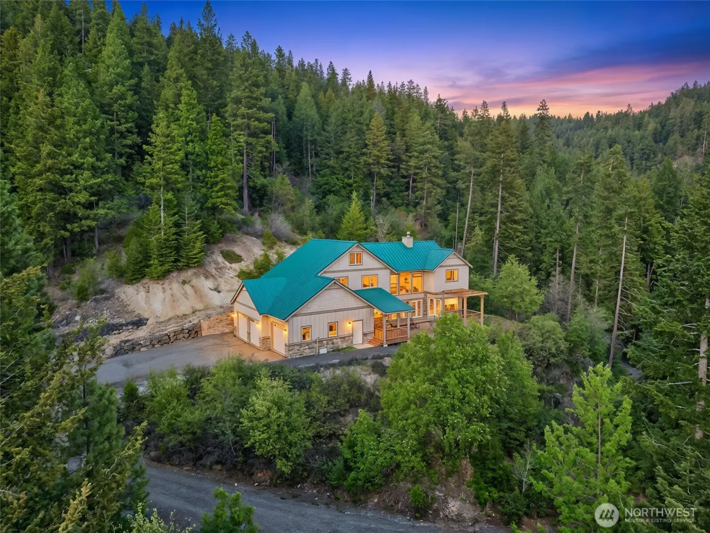 A two-story home with a green metal roof nestled in a dense pine forest at sunset, featuring a driveway and warm lighting.