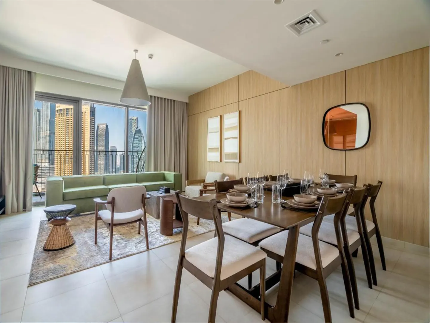 Bright apartment interior with a dining table set for a meal, a green sofa, and a city view through large windows.