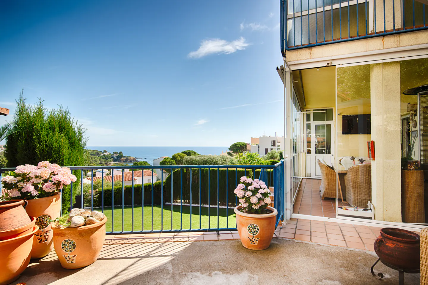 Sunny terrace with potted flowers, blue railings, and a scenic view of the Mediterranean sea under a clear blue sky.