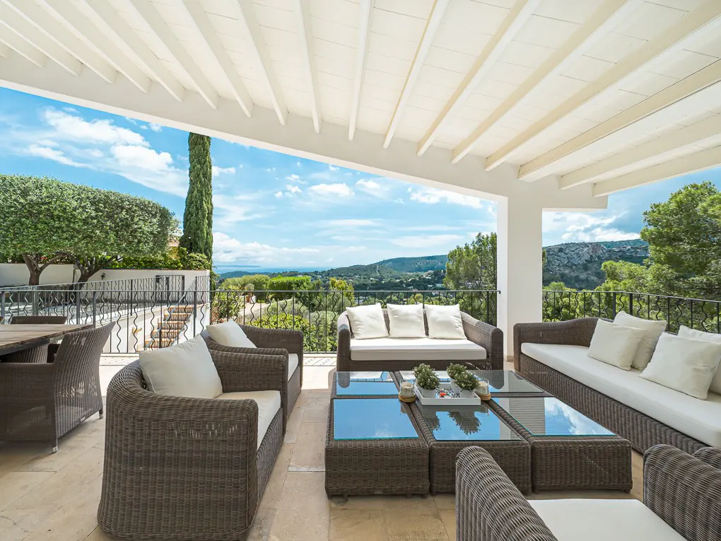 Outdoor patio with wicker furniture, white cushions, and glass tables. A scenic view of green hills and a blue sky is in the background.