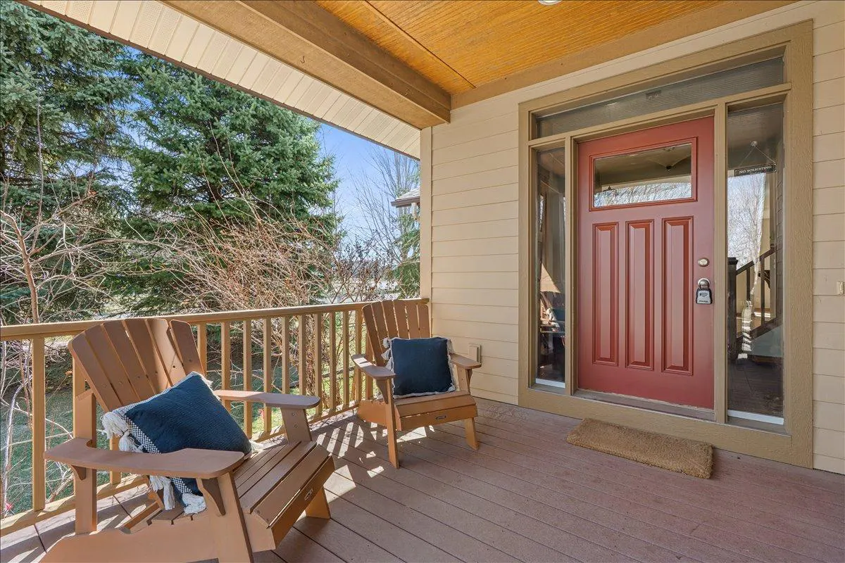 Covered porch with two wooden Adirondack chairs with blue pillows, a red front door with sidelights, and a view of trees.