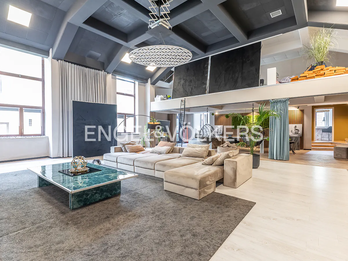 A modern living room with a large beige sectional sofa, a green marble coffee table, and a gray rug. The room has high ceilings and large windows.