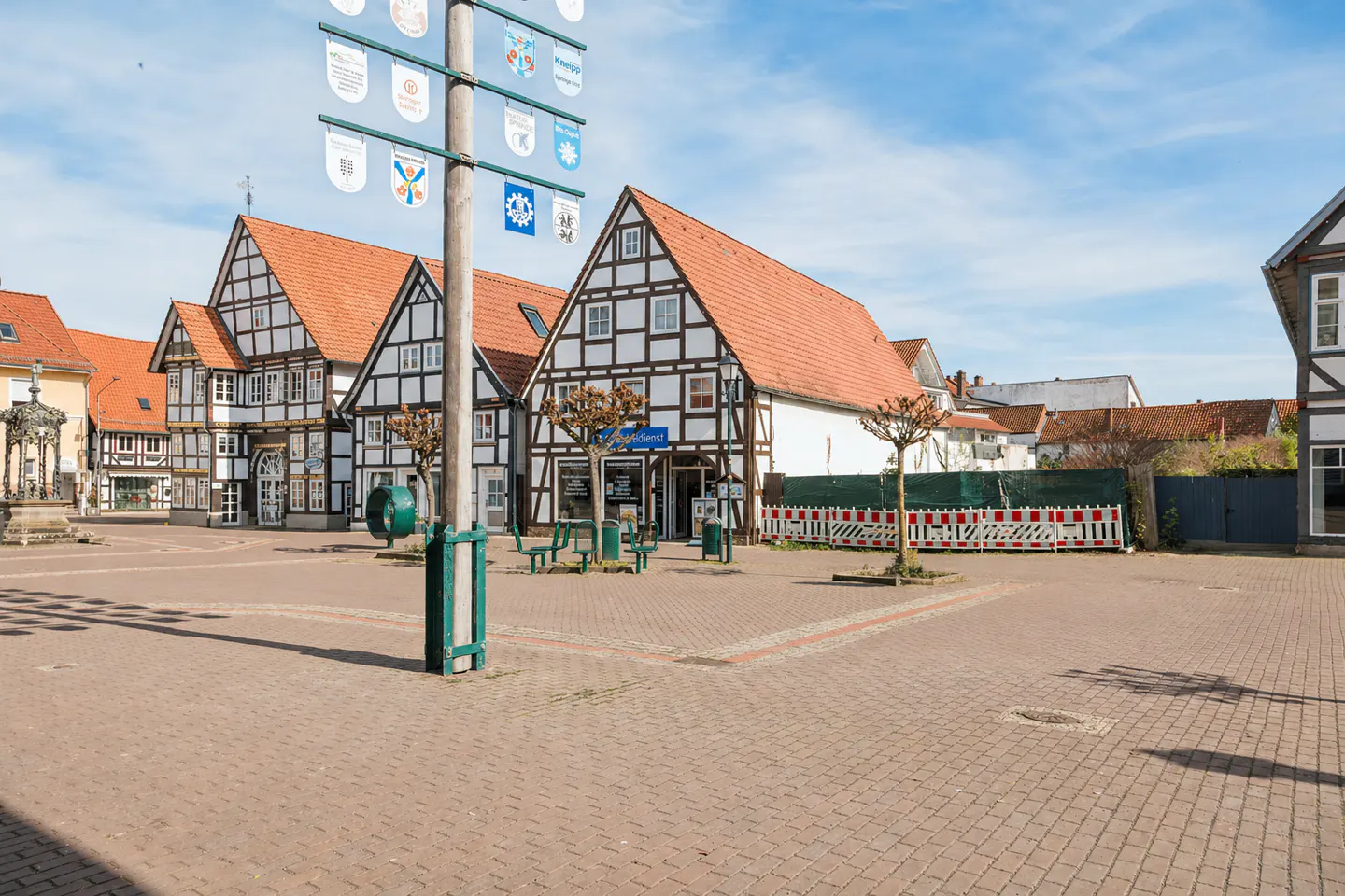 Town square with half-timbered houses, red tile roofs, and a brick-paved plaza under a blue sky. A tall pole with banners stands in the foreground.