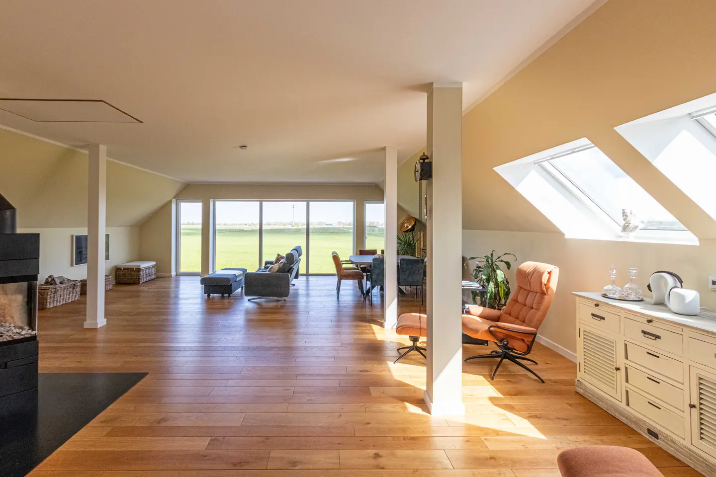 Bright, open living space with wood floors, fireplace, and large windows overlooking a green field. Skylights and a cream-colored cabinet add to the room's light.
