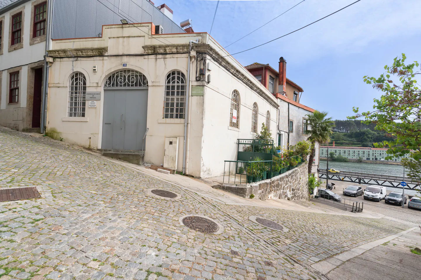 Exterior of a white building with arched windows on a cobblestone street. Cars are parked near a river in the background.