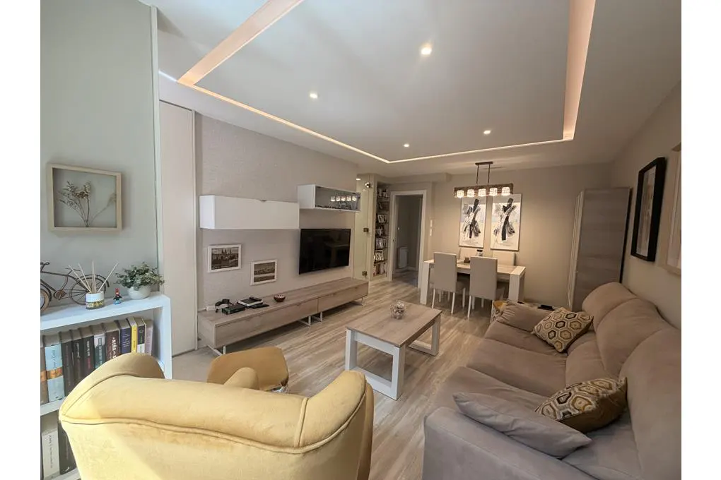 A modern living room with a beige sofa, yellow armchair, and dining area. A TV and decorative items adorn the wall unit.