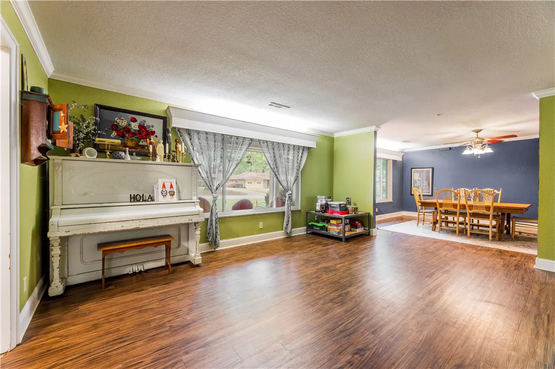 A wide shot of a living room with a piano, wood floors, and green walls. A dining room is visible in the background.