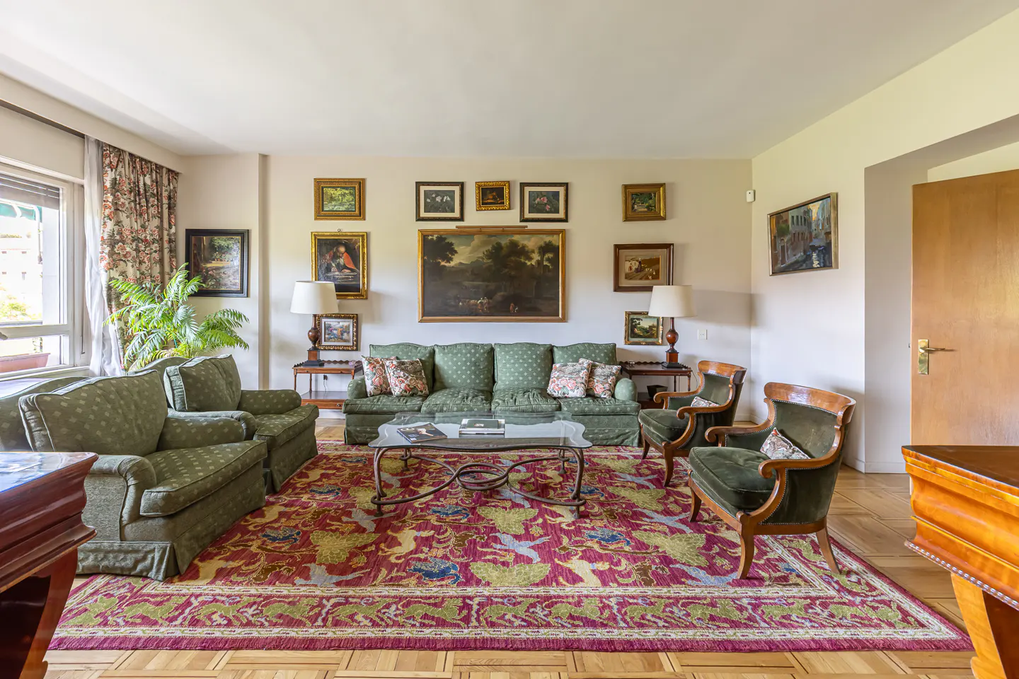Living room with green sofas, chairs, and a red patterned rug. Art hangs on the wall above the sofa. A glass coffee table sits in the center.