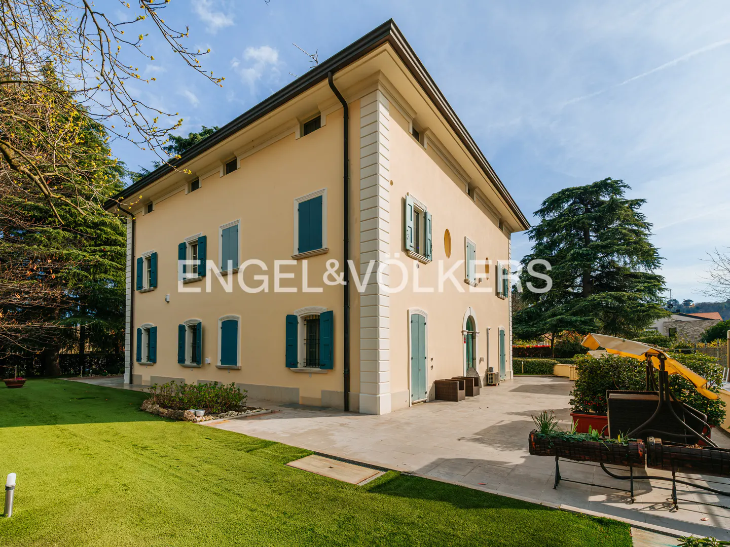 Exterior view of a two-story beige house with blue shutters, a green lawn, and the Engel & Völkers logo.