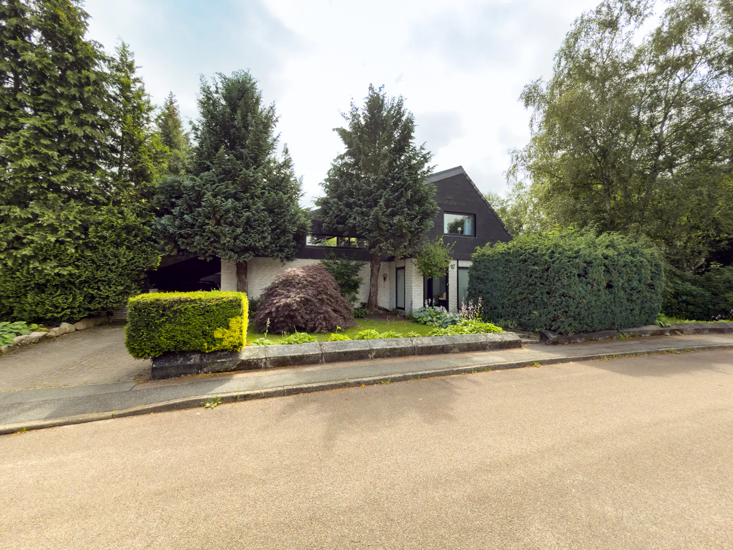 A modern house with a black roof and white walls is surrounded by green trees and bushes.