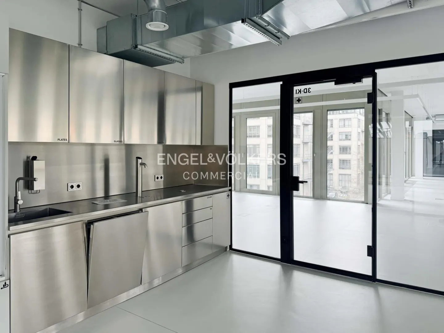 Modern office space with stainless steel kitchen, sink, and cabinets. Black framed glass doors lead to a bright, open area. Engel & Volkers logo visible.