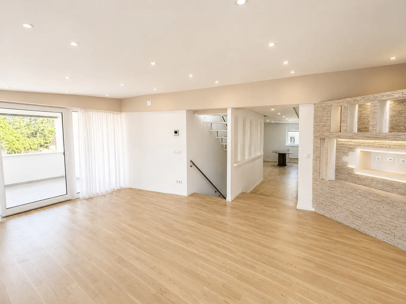 Bright, empty living room with light wood floors, white walls, and recessed lighting. A staircase and balcony door are visible.