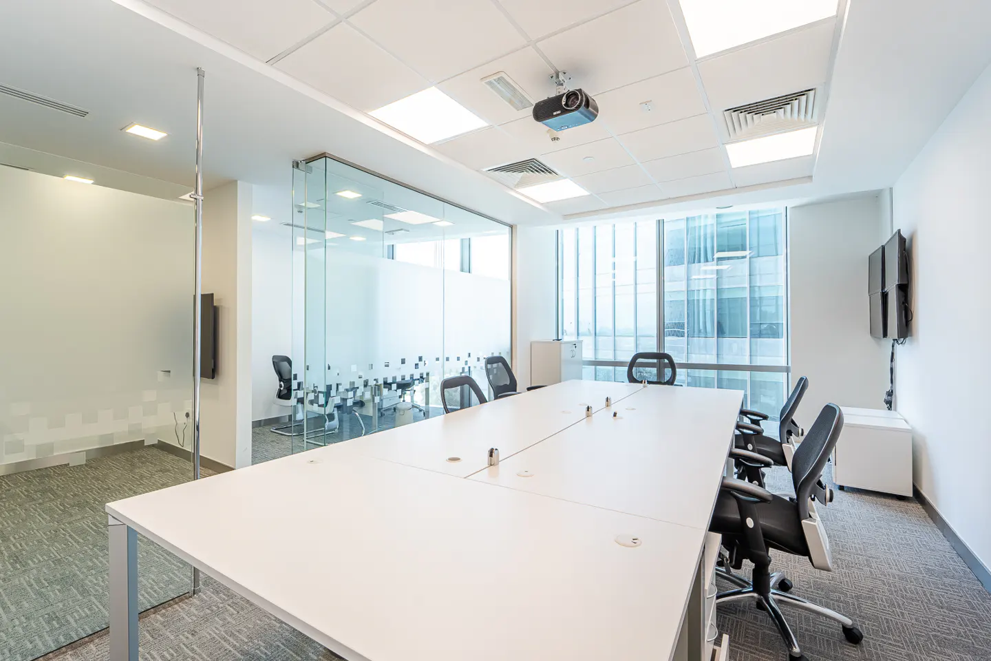 Bright, modern office conference room with a long white table, black chairs, glass walls, and a projector on the ceiling.