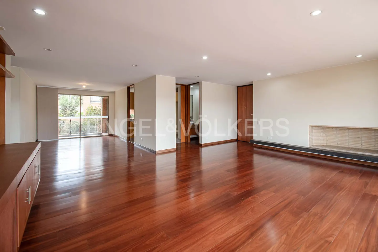 Spacious living room with shiny hardwood floors, white walls, and recessed lighting. A fireplace and balcony doors add natural light.