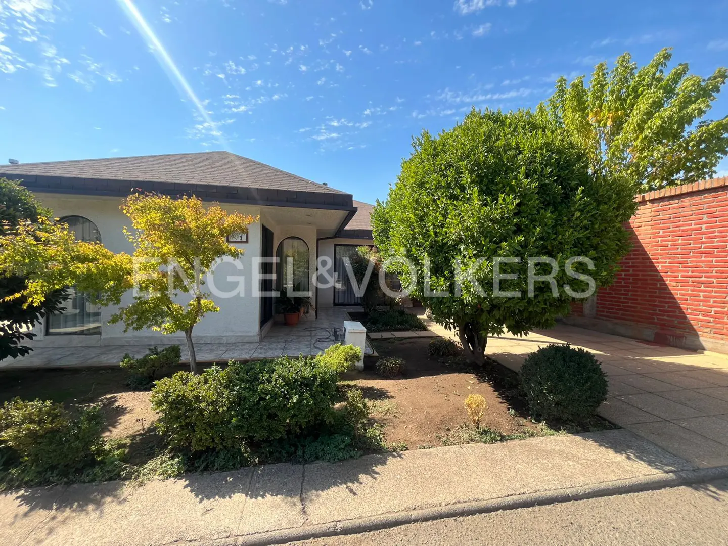Exterior of a one-story white house with a brown roof, green trees, and a brick wall under a blue sky.