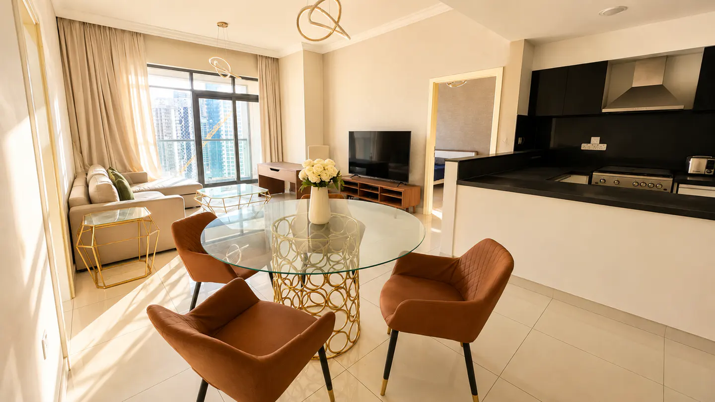 A modern apartment interior with a glass table, brown chairs, and a beige sofa near a window with city views.