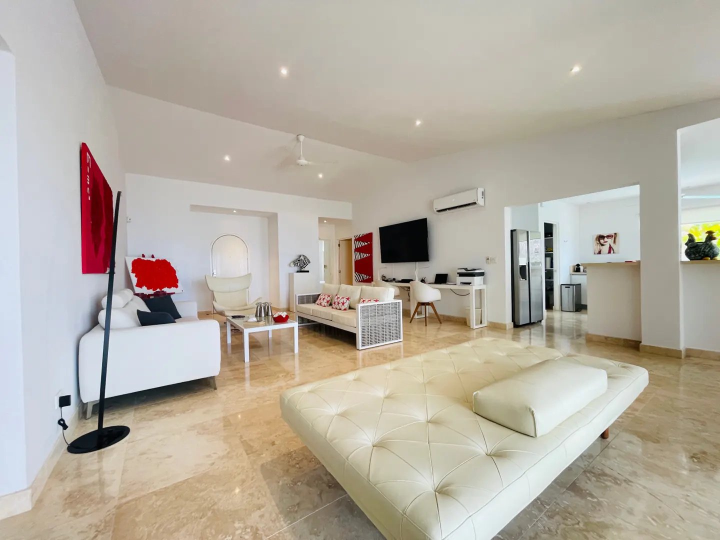 Bright, open-concept living space with white sofas, a desk, and a kitchen visible in the background. The floor is light brown marble.
