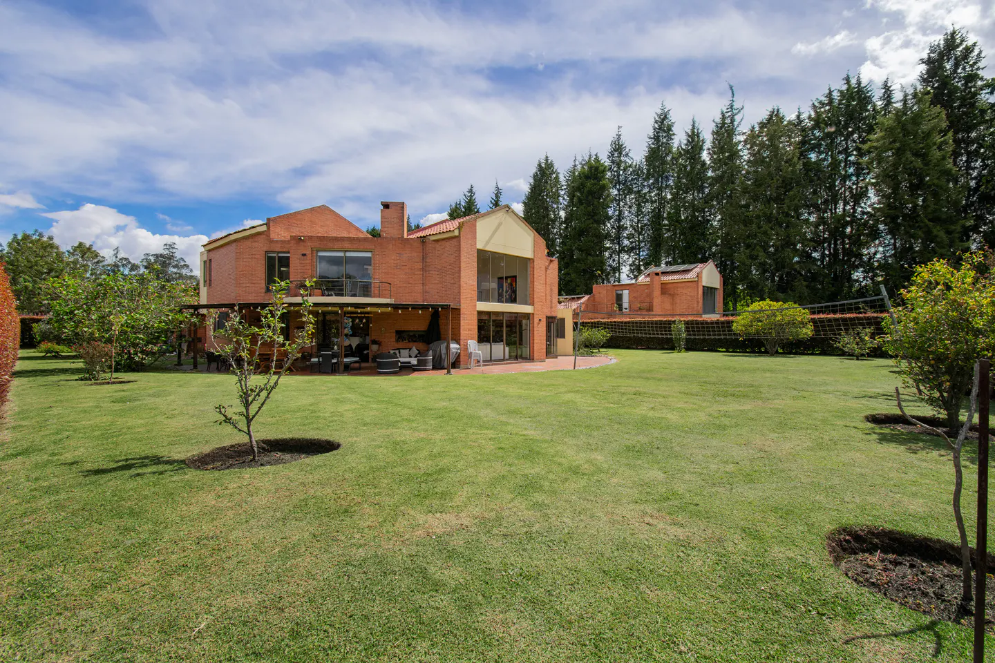Brick house with a large green lawn, trees, and blue sky with white clouds. The house has a patio with furniture.