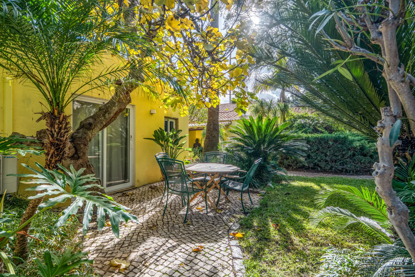 Outdoor patio with a round table and four chairs on a stone patio, surrounded by lush greenery and a yellow building.
