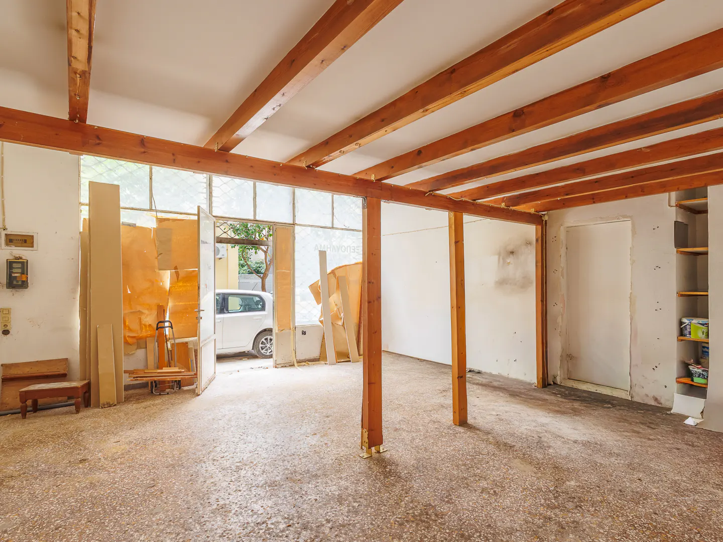 Empty room under renovation with exposed wooden ceiling beams, concrete floor, white walls, and an open door to a street.