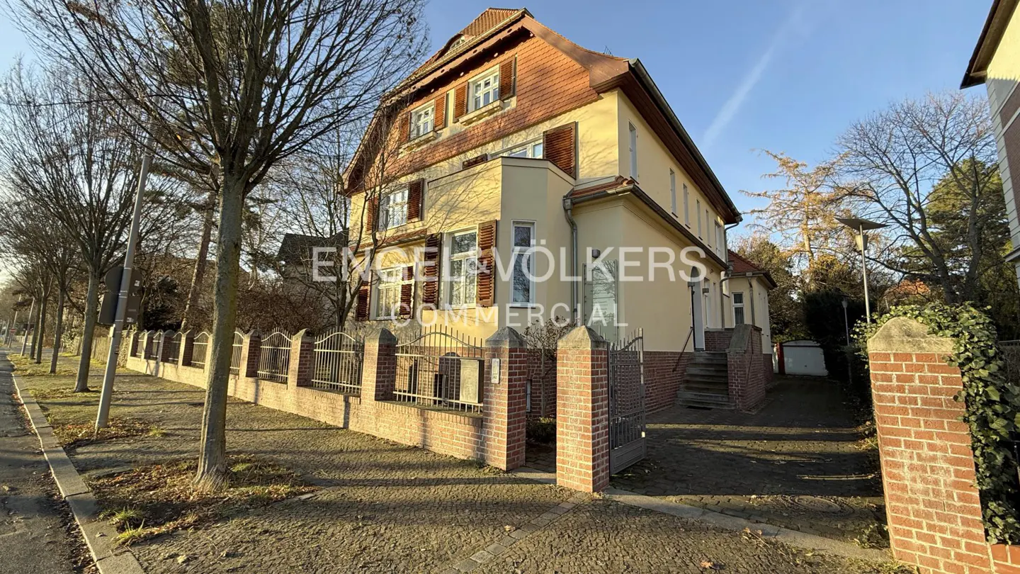 Two-story yellow house with a red tile roof and brown shutters, behind a brick and iron fence. Trees line the street.