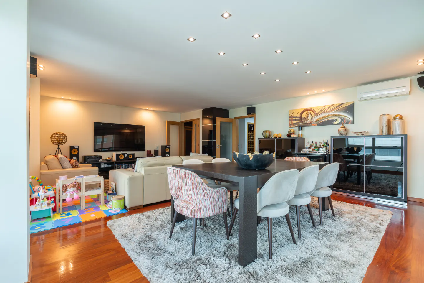 Open-concept living and dining area with hardwood floors, a gray rug, and a dark wood dining table with six chairs. A TV and sofas are in the background.