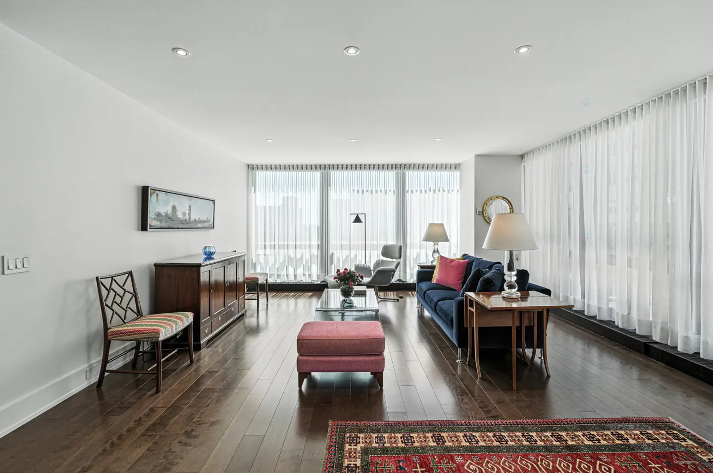 Bright living room with hardwood floors, white walls, and large windows with sheer white curtains. A blue sofa, pink ottoman, and patterned rug add color.