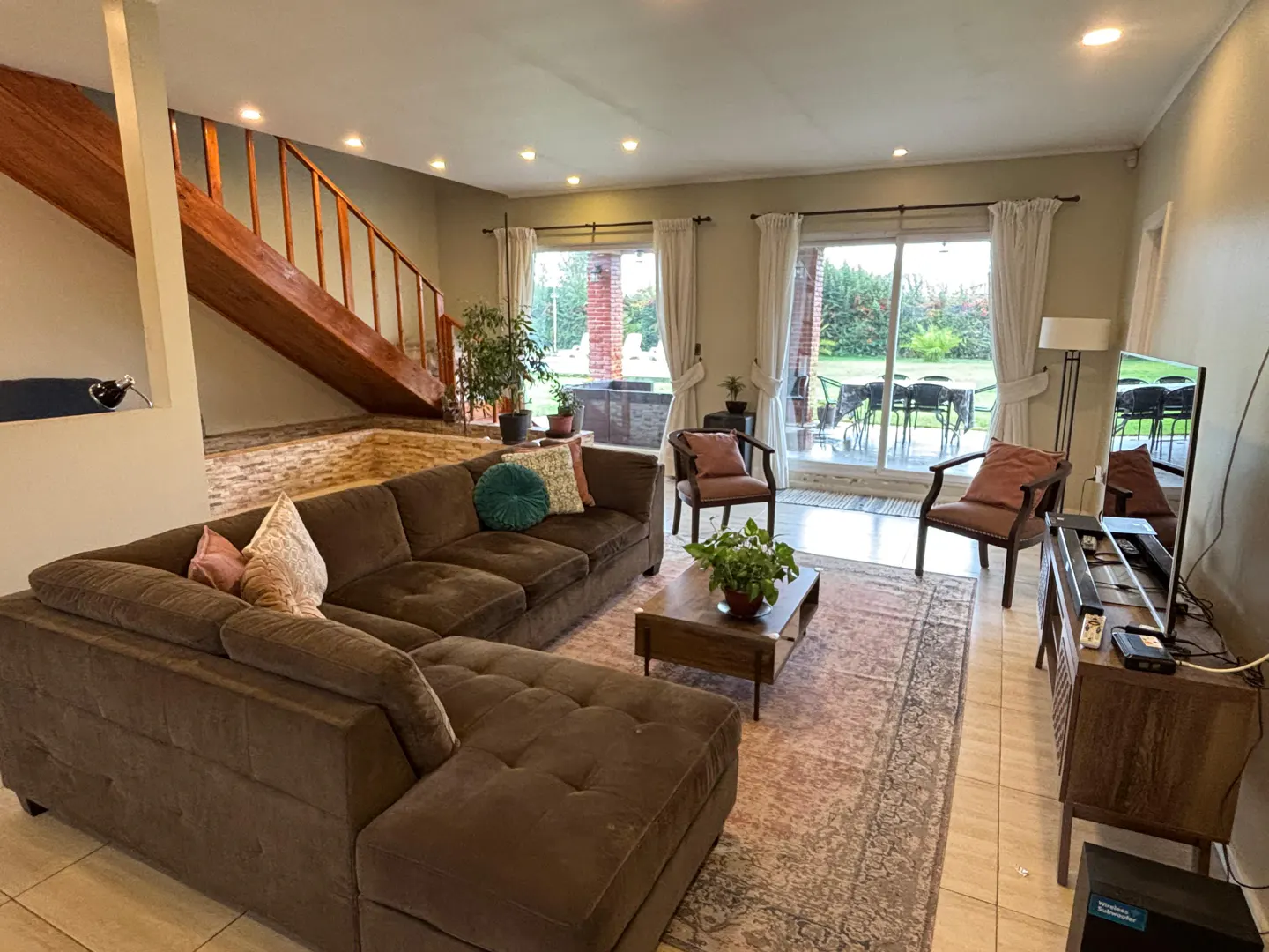 A living room with a brown sectional sofa, wooden stairs, and a view of the backyard through sliding glass doors.