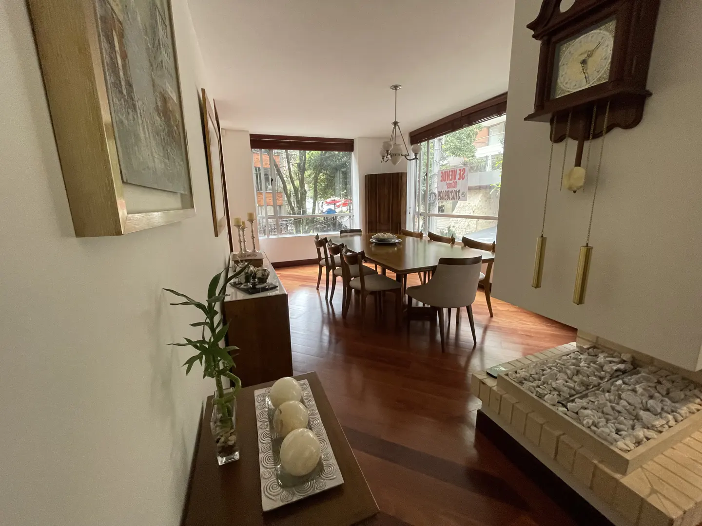 A dining room with hardwood floors, a wooden table with chairs, and a grandfather clock on the wall. A window overlooks a city street.