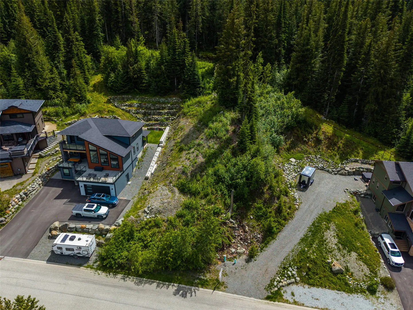 Aerial view of modern homes nestled in a lush, forested mountain landscape with paved driveways and parked vehicles.