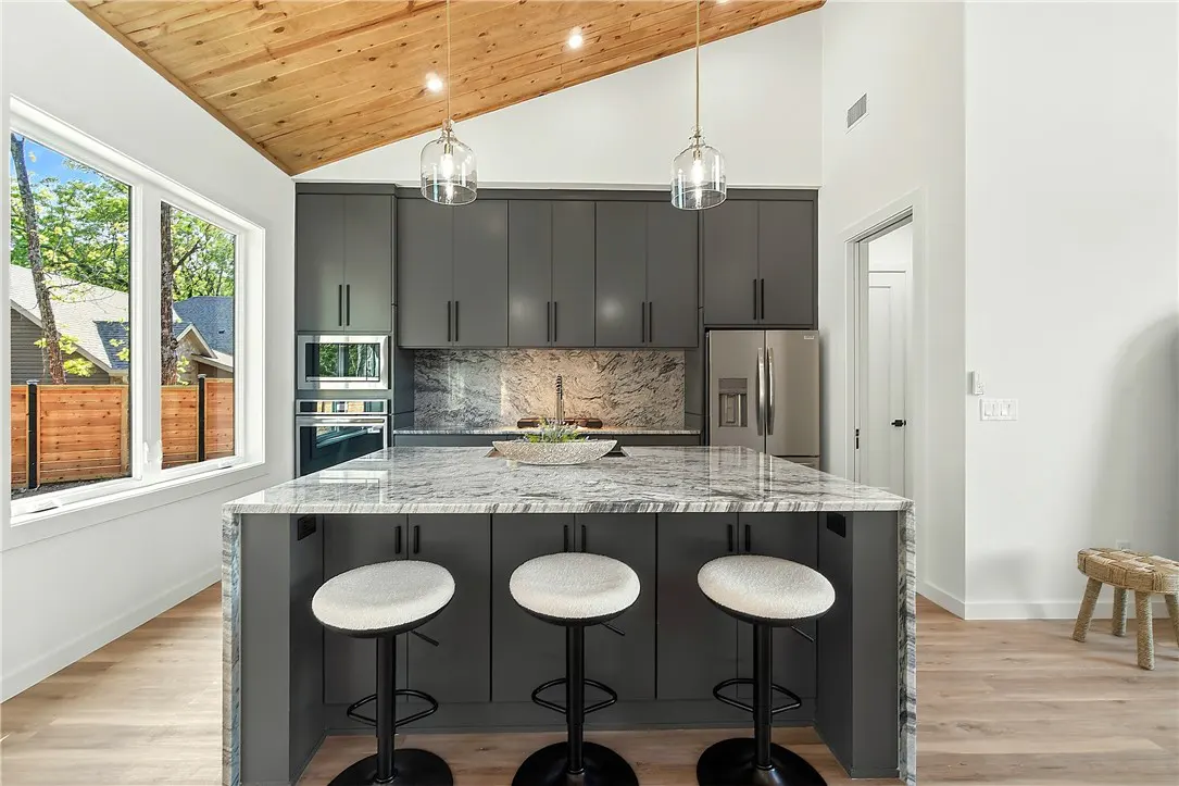 Modern kitchen with gray cabinets, marble countertops, and a wood ceiling. Three stools line the island. A large window offers a view of trees.