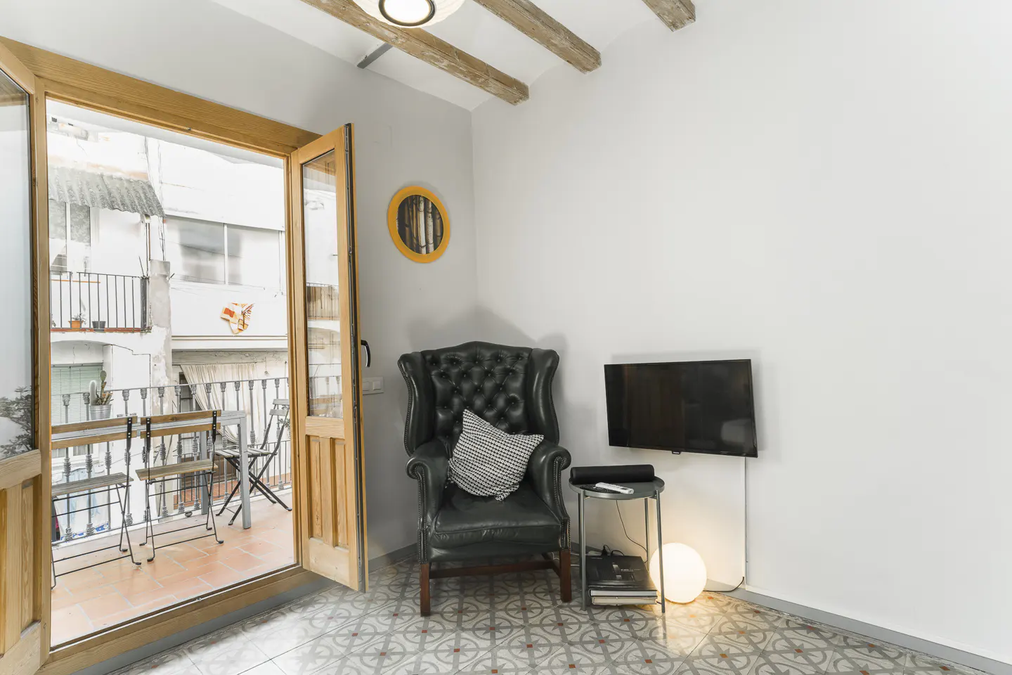 A bright living room with a black leather armchair, a TV, and an open door to a balcony with a table and chairs.