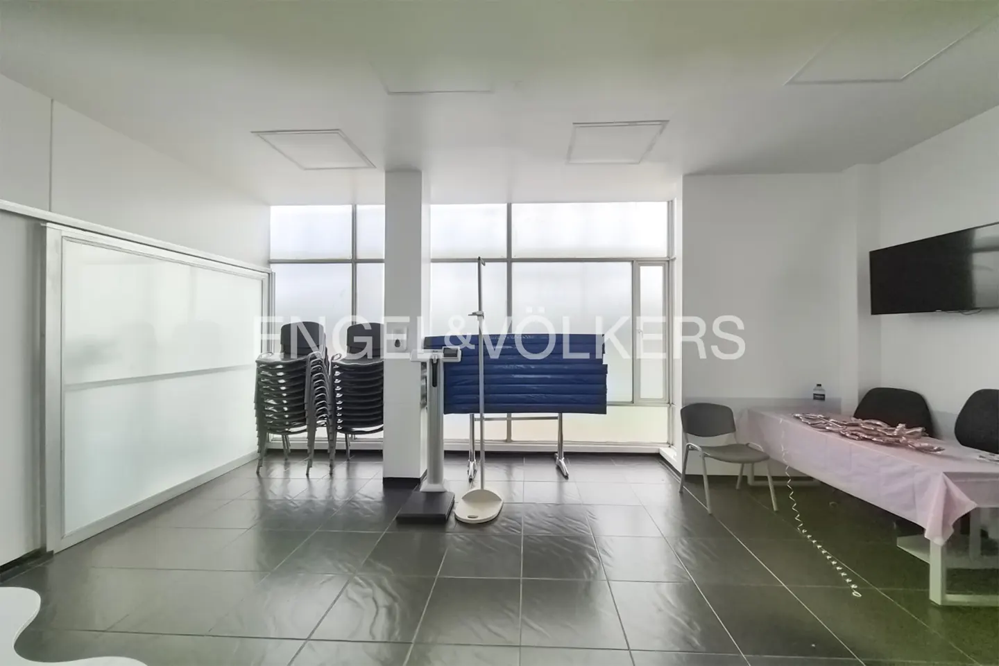 Bright room with black tile floor, white walls, and large windows. Stacked chairs, scale, and table with pink tablecloth are visible.