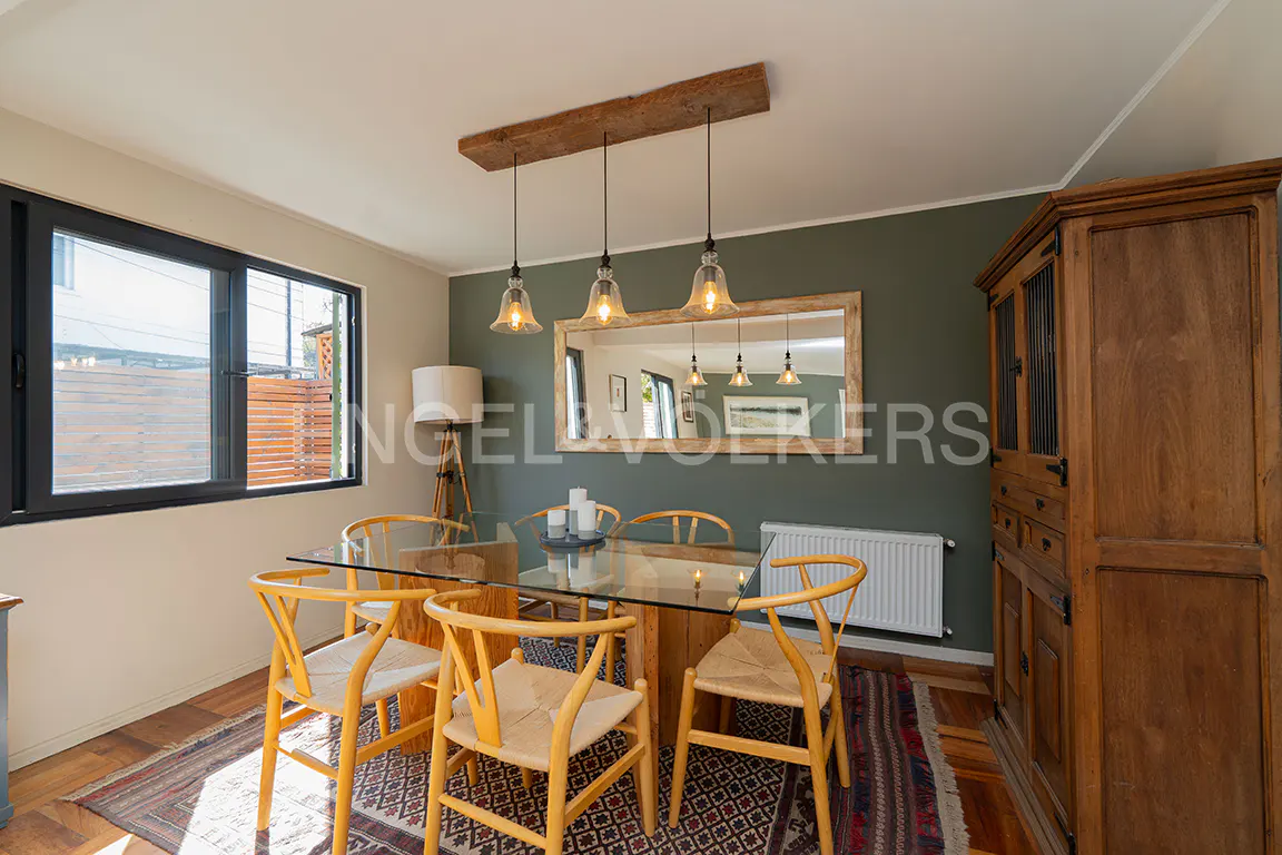 Dining room with glass table, wooden chairs, and a rustic wooden light fixture. A large wooden cabinet stands against a dark green wall.