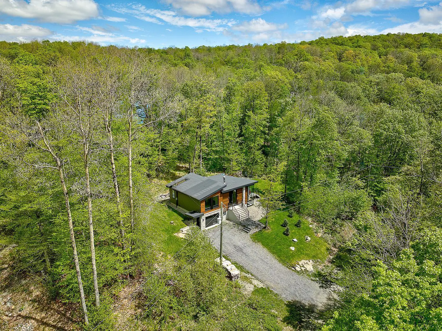 Aerial view of a modern home with a dark roof nestled in a lush, green forest with a gravel driveway.