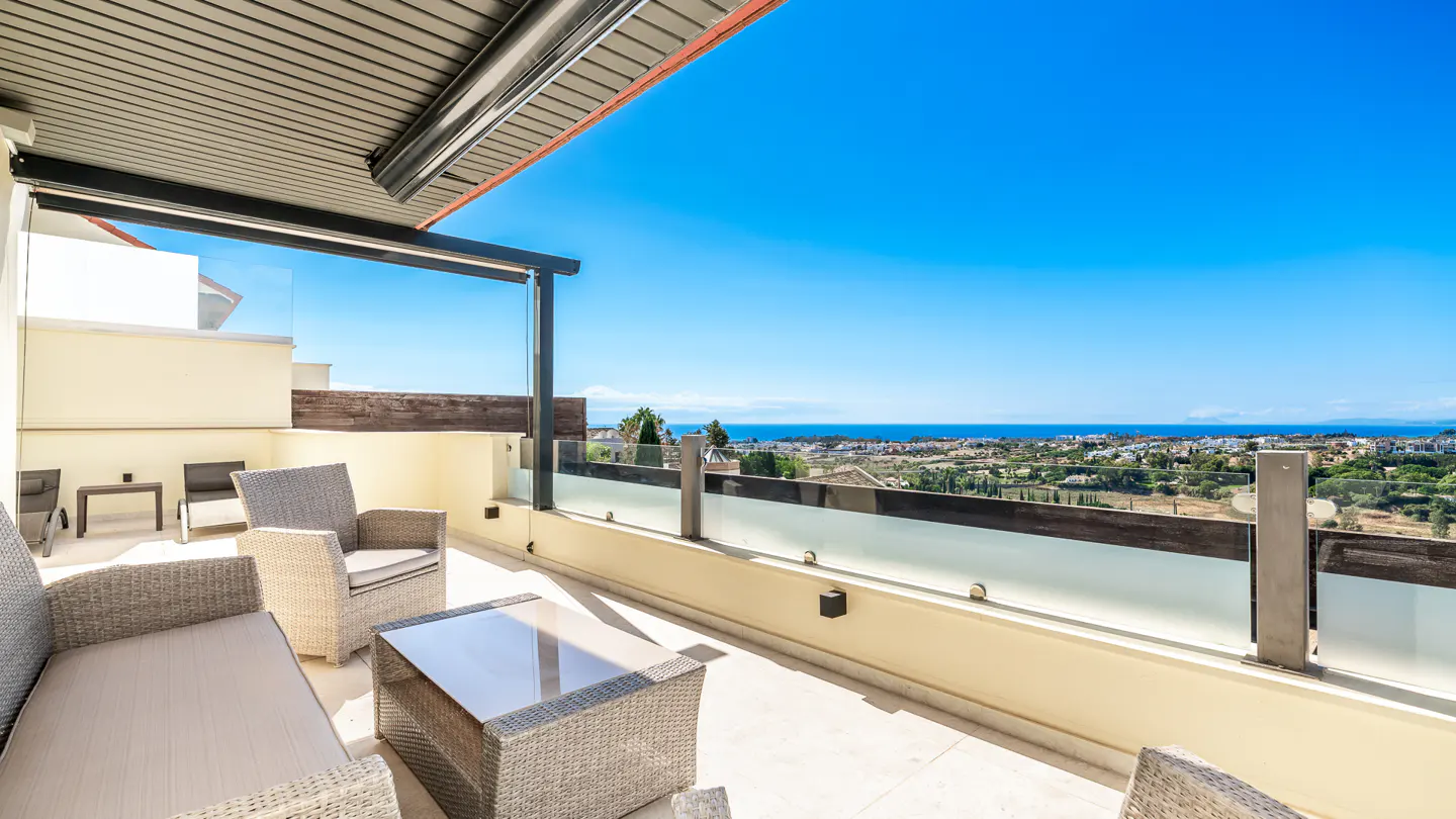 Outdoor patio with wicker furniture, glass railing, and a view of the ocean and blue sky.