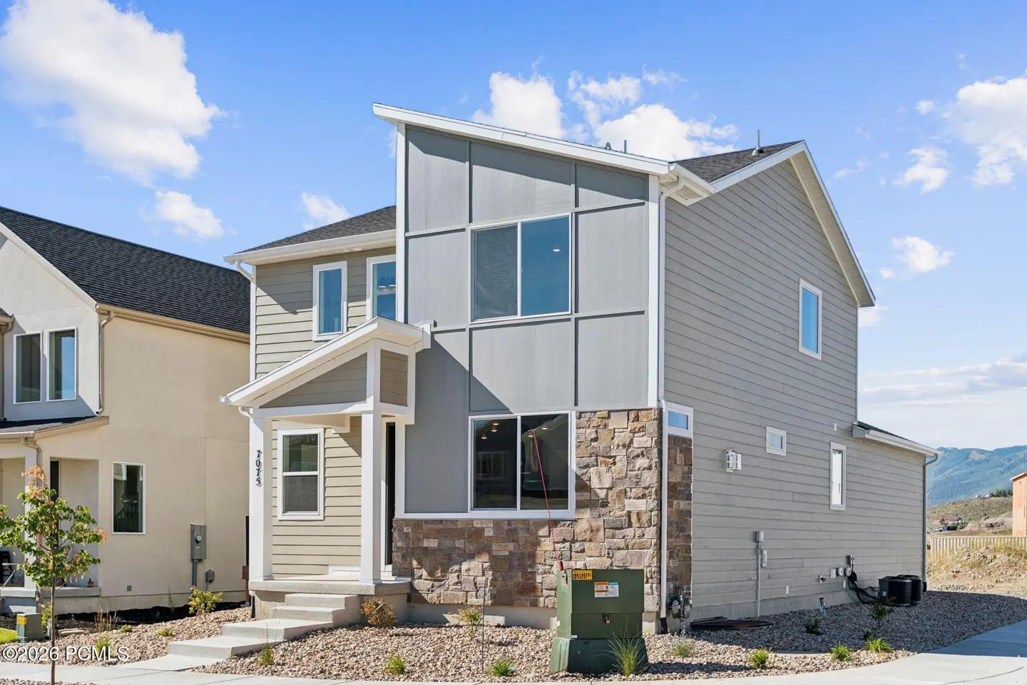 Modern two-story home with gray siding, stone accents, and a slanted roof under a bright blue sky with scattered clouds.
