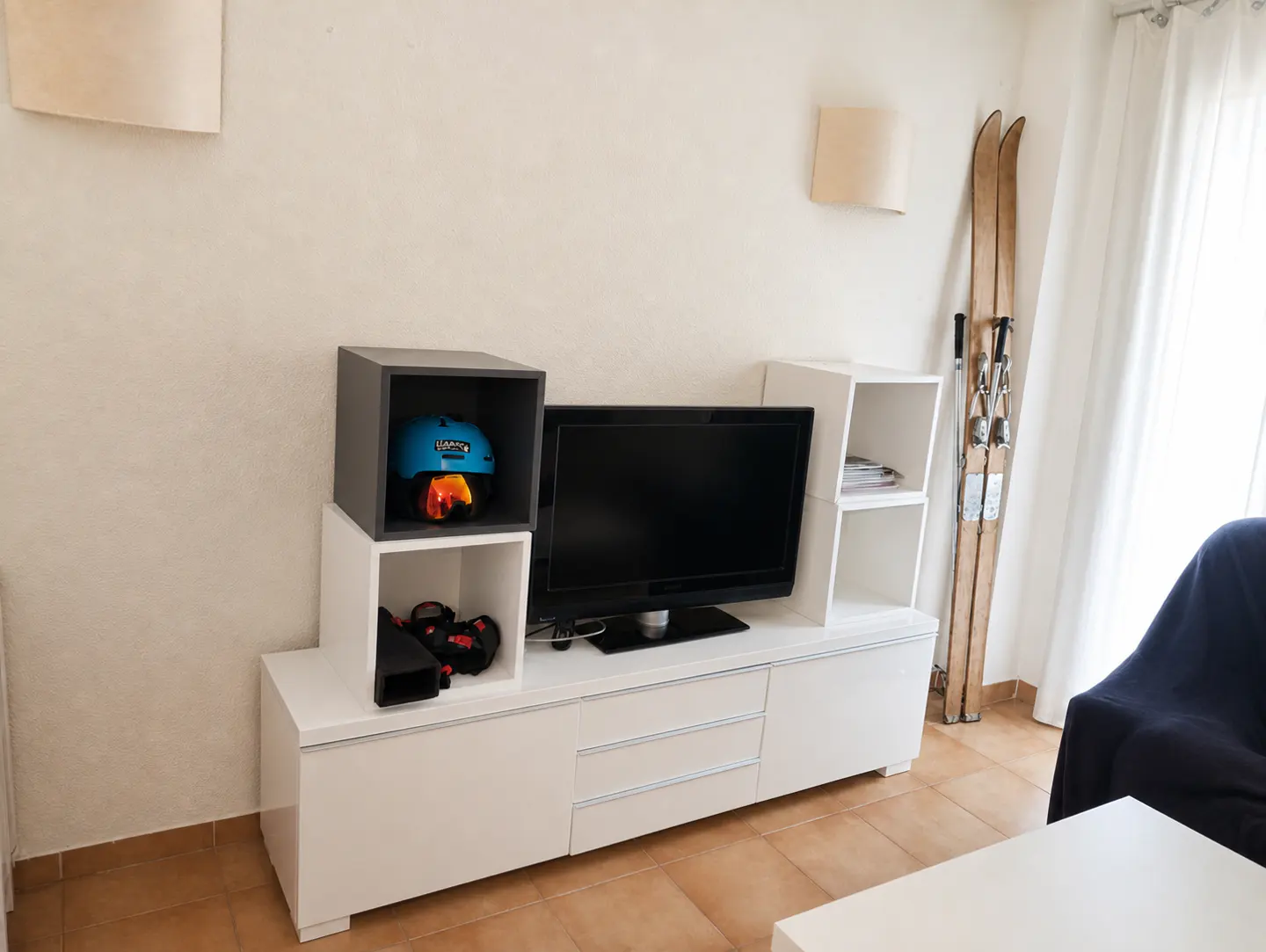 Modern white media console with a TV, a blue ski helmet in a cubby, and vintage wooden skis leaning against the wall.