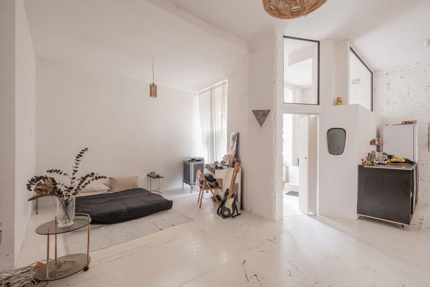 Bright, open studio apartment with white walls and floors. A bed, desk, and kitchenette are visible. A guitar leans against the desk.