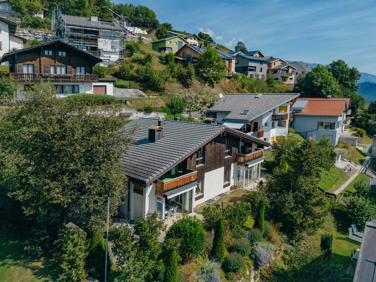 A hillside neighborhood with houses of varying styles and colors, surrounded by lush greenery under a blue sky.