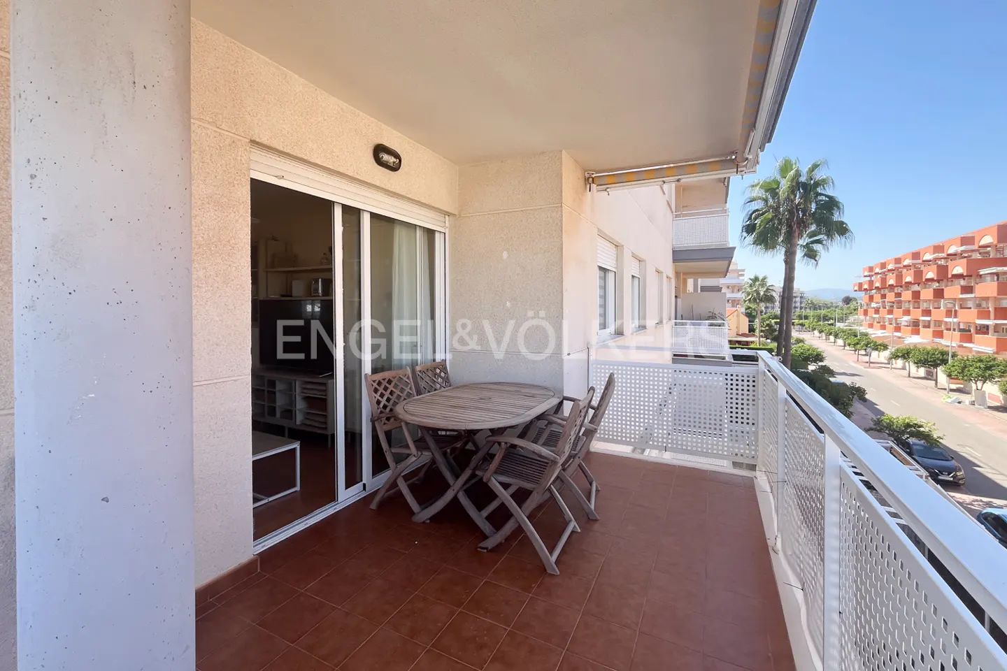 Balcony view with a round wooden table and chairs on brown tile flooring, overlooking a street with palm trees and buildings.