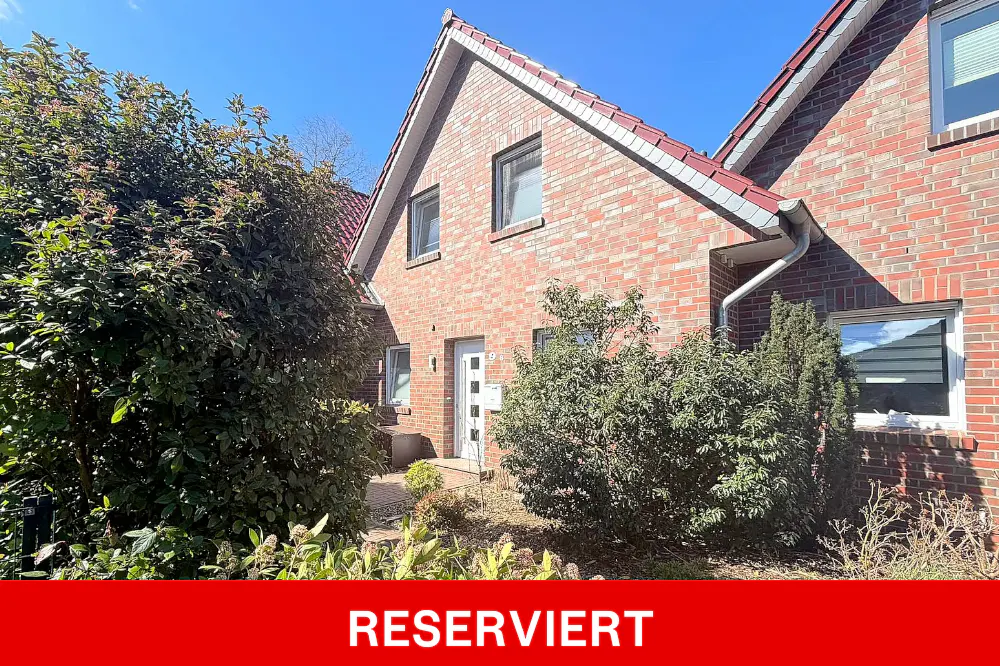 Exterior view of a two-story brick house with a red tile roof and white door, surrounded by green bushes. "RESERVIERT" is written in white on a red banner.